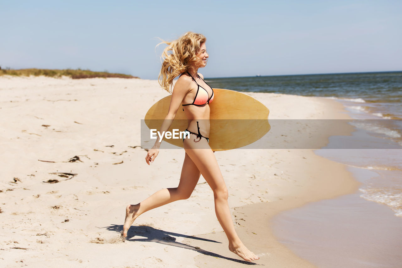 Woman in bikini carrying surfboard while running on shore at beach