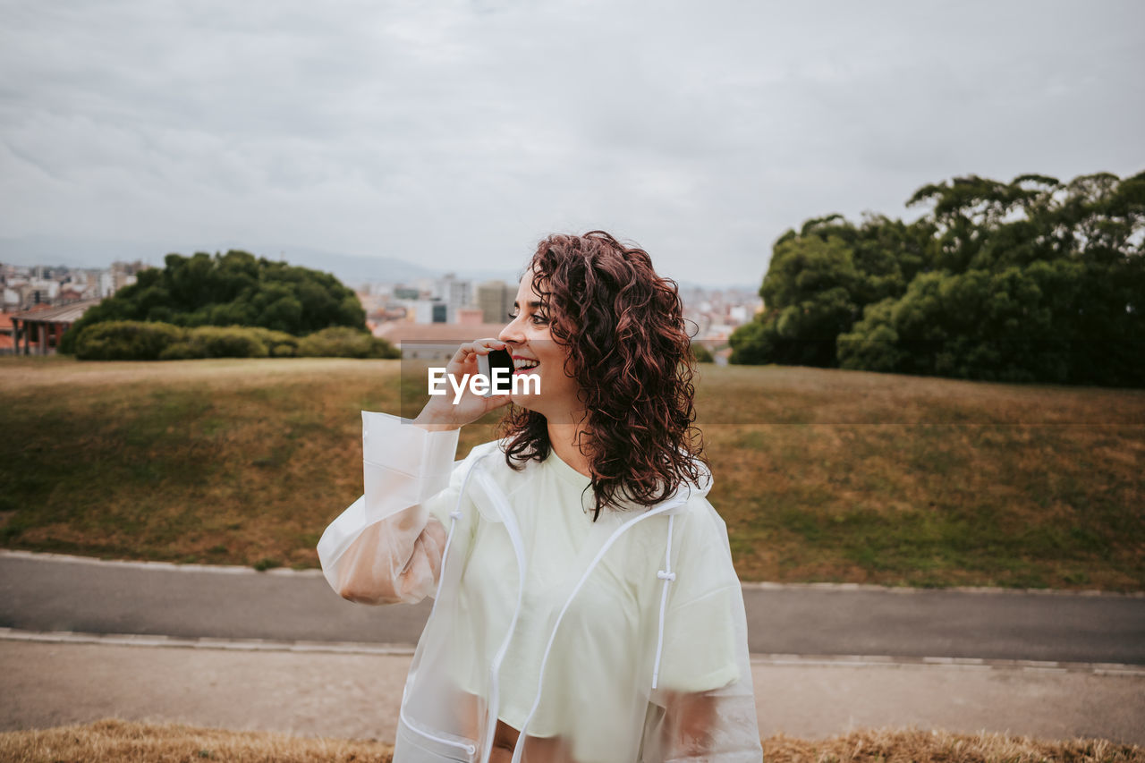 Smiling woman talking on phone looking away standing against sky