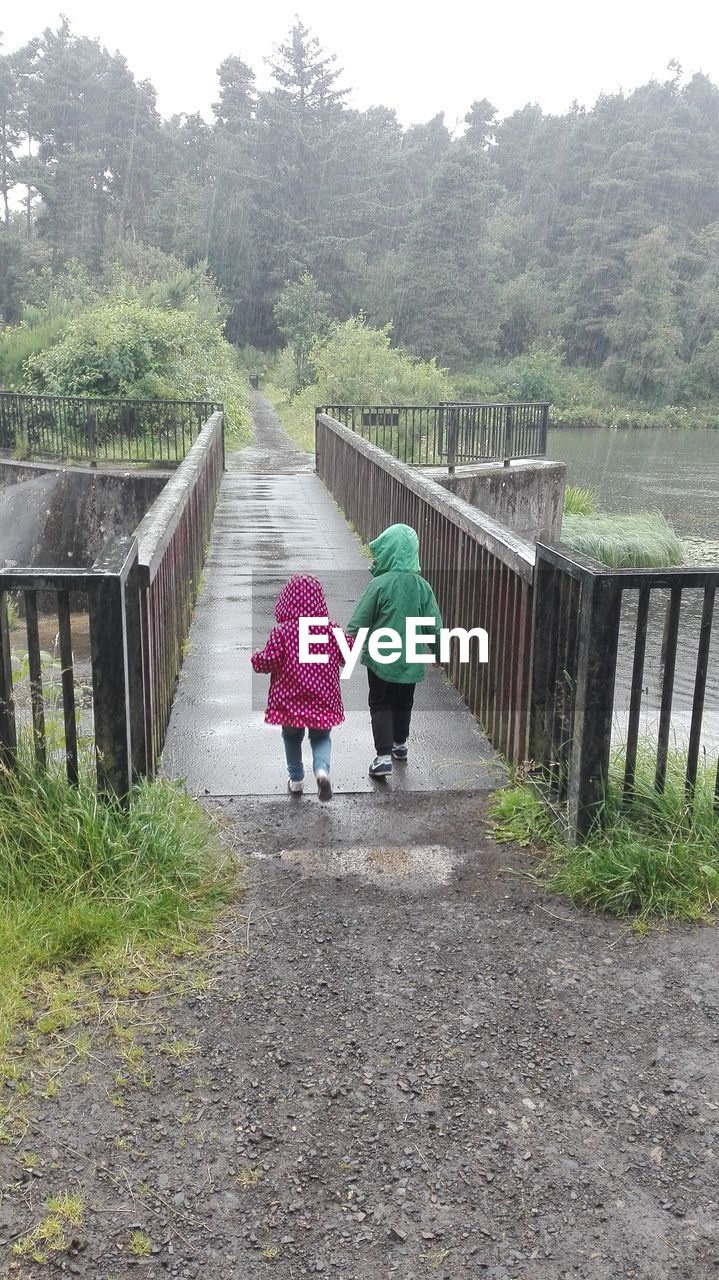 Rear view of siblings wearing raincoats while walking on footbridge over river during rainfall