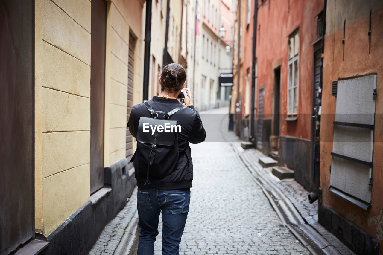 Rear view of man photographing through camera while standing on cobbled street in alley