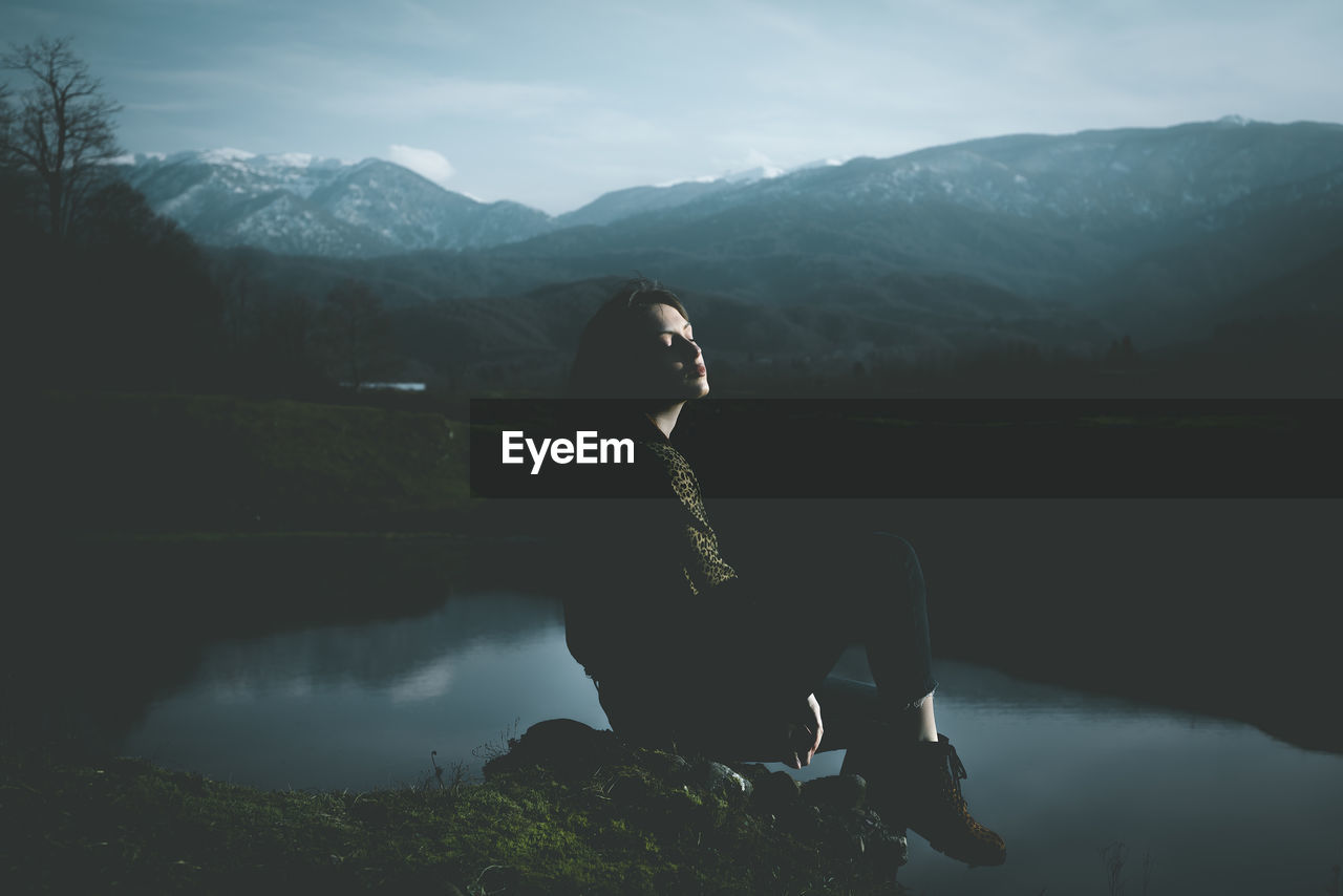 Woman sitting by lake against mountains