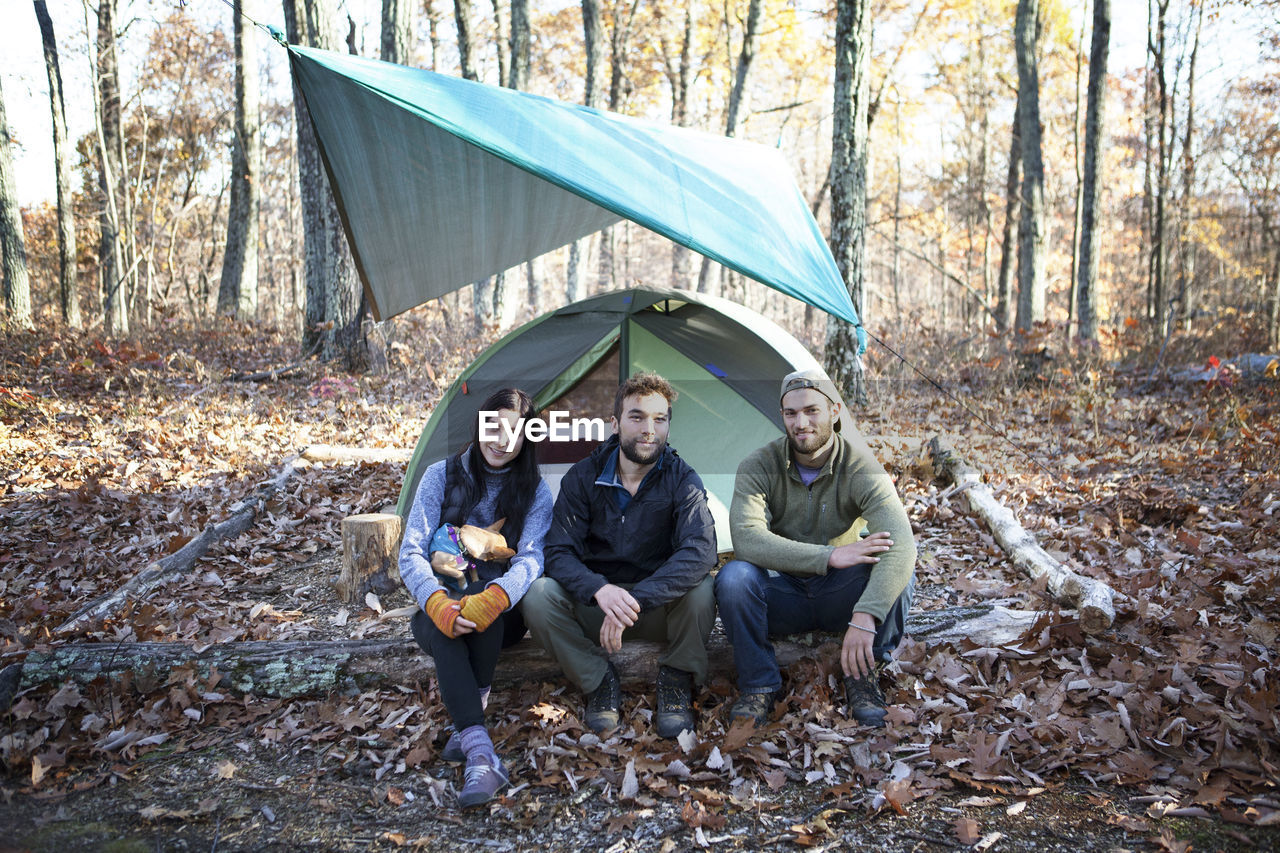 Portrait of friends sitting on fallen tree trunk by tent in forest