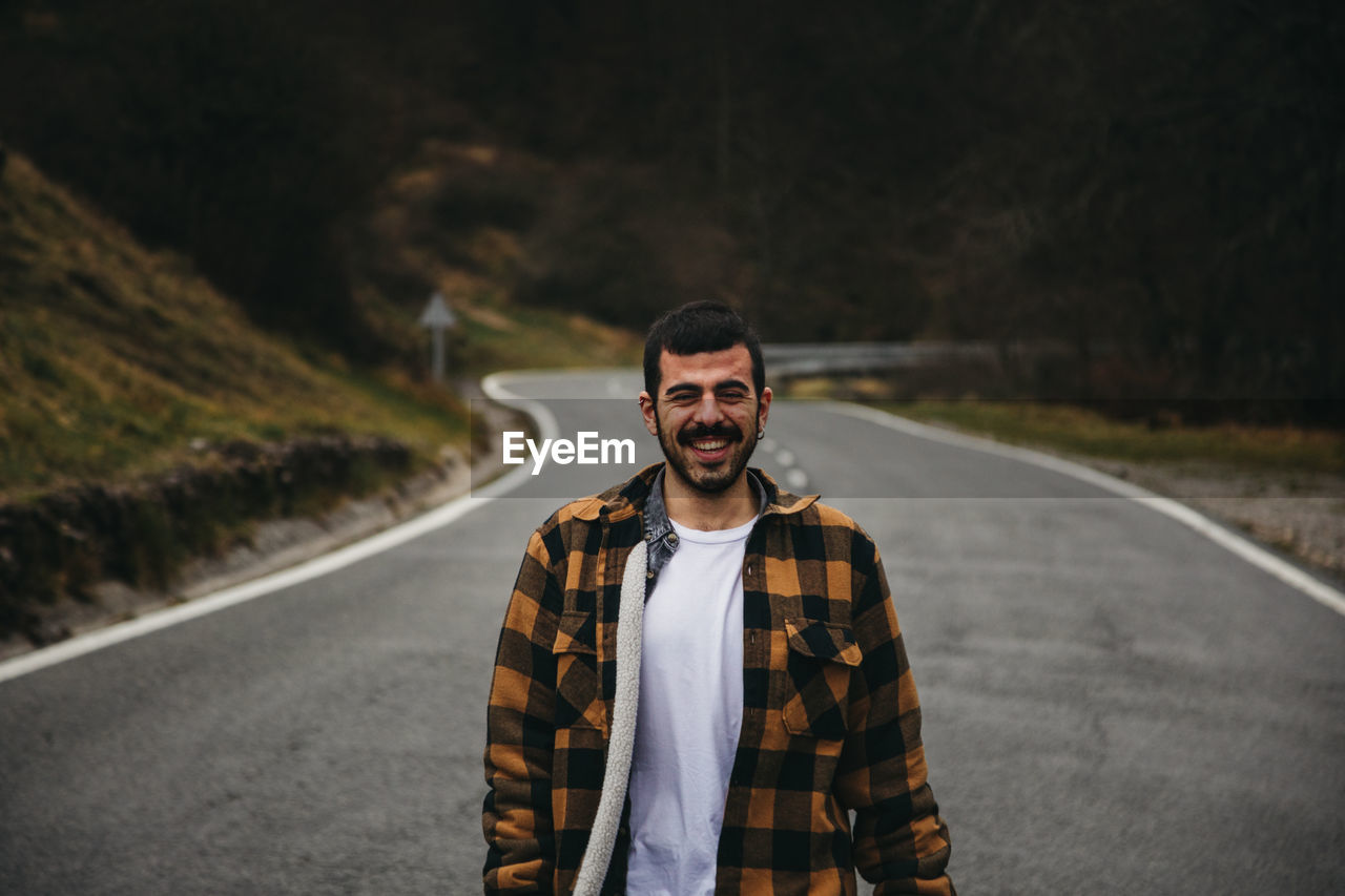 Bearded black haired cheerful man in casual wear smiling and looking at camera while standing on empty road in autumn countryside