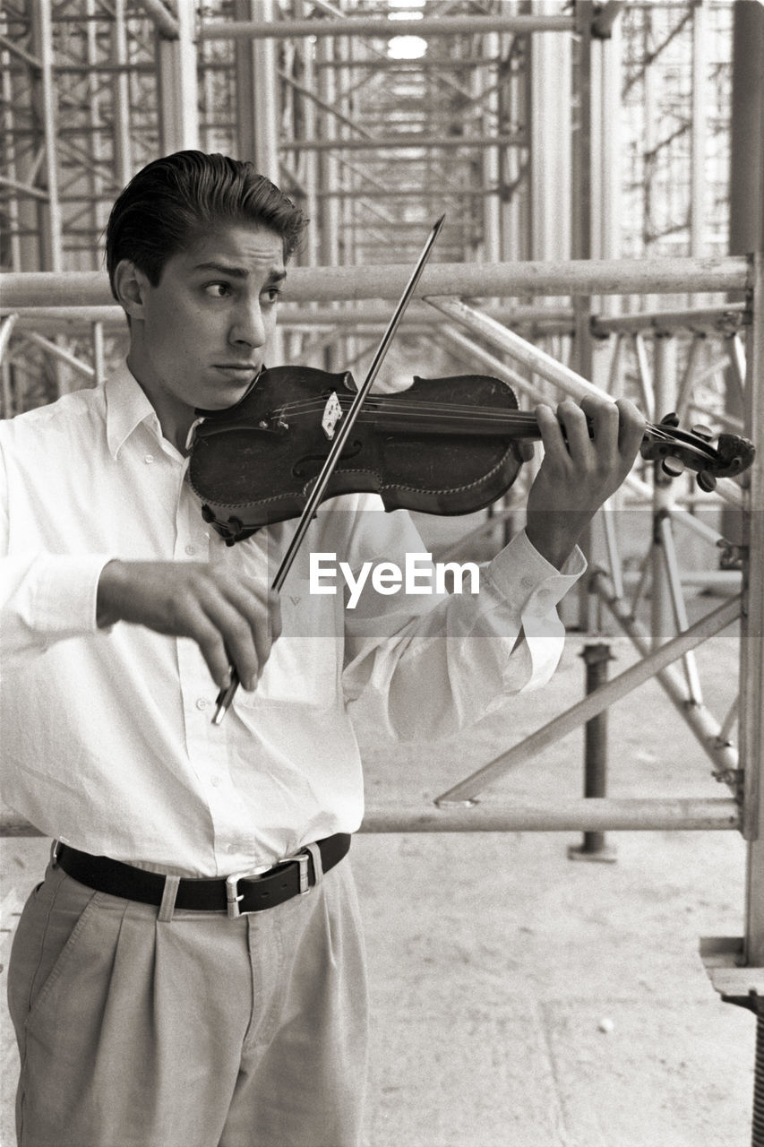 Man playing violin while standing on footpath against scaffolding