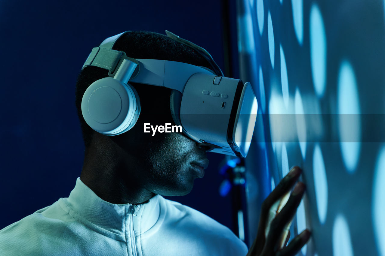 Side view of concentrated young african american in white t shirt and modern vr goggles pointing and touching screen while interacting with virtual object in dark studio with neon illumination