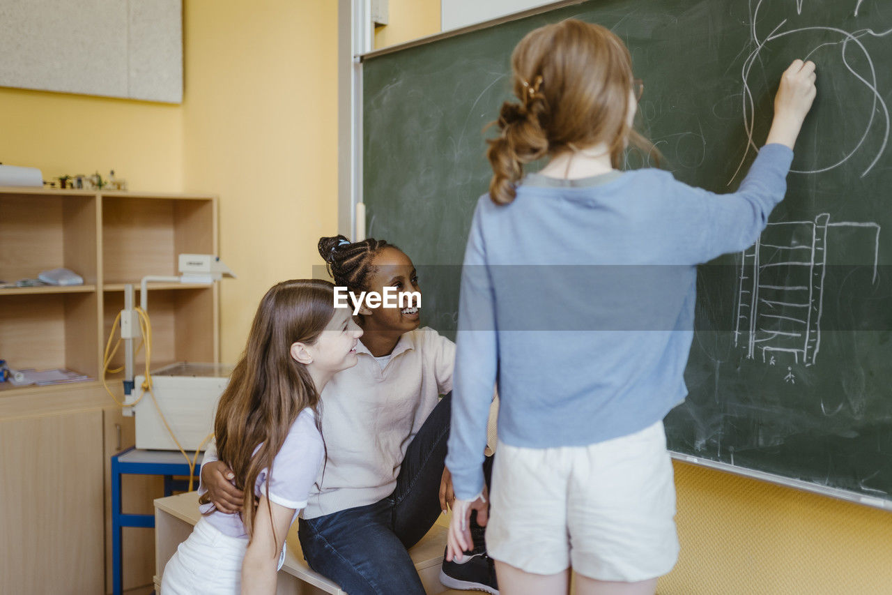 Girls having fun with female friend drawing on chalkboard in classroom