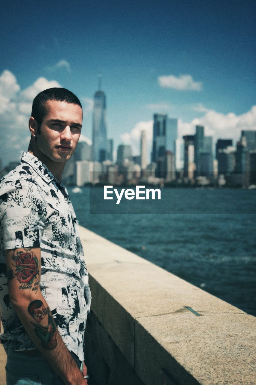 Portrait of young man standing by river against sky in city