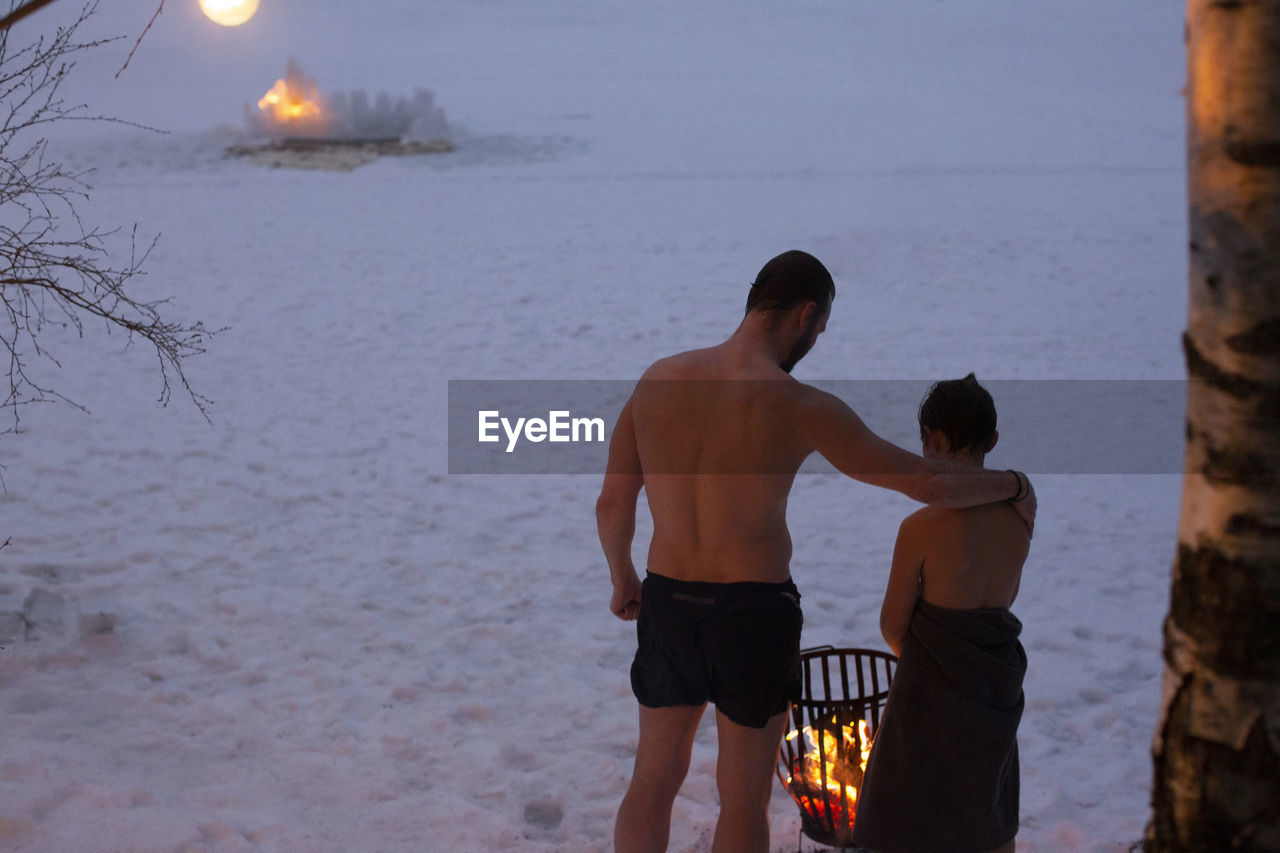 Rear view of shirtless father standing with son by bonfire at night