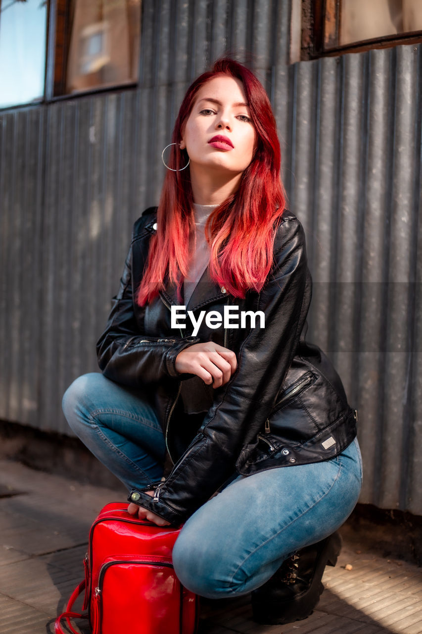 red, one person, women, adult, young adult, sitting, portrait, clothing, leather, fashion, hairstyle, casual clothing, leather jacket, architecture, lifestyles, looking at camera, jacket, city, full length, long hair, female, person, redhead, photo shoot, front view, leisure activity, footwear, city life, individuality, arts culture and entertainment, jeans, emotion, relaxation, dyed red hair, cool attitude, luggage and bags, looking