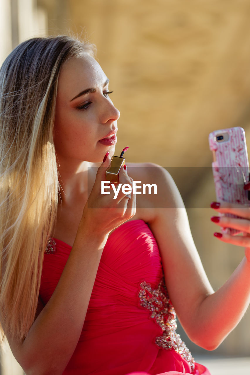 Young woman in pink evening gown applying lipstick while looking at smart phone outdoors