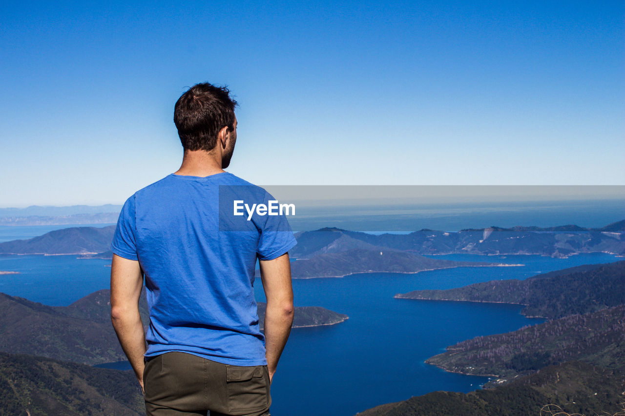 Rear view of man looking at sea while standing on mountain against blue sky