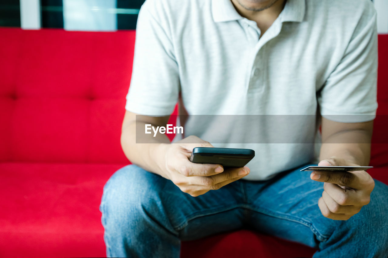 Midsection of man using mobile phone and holding credit card while sitting on sofa