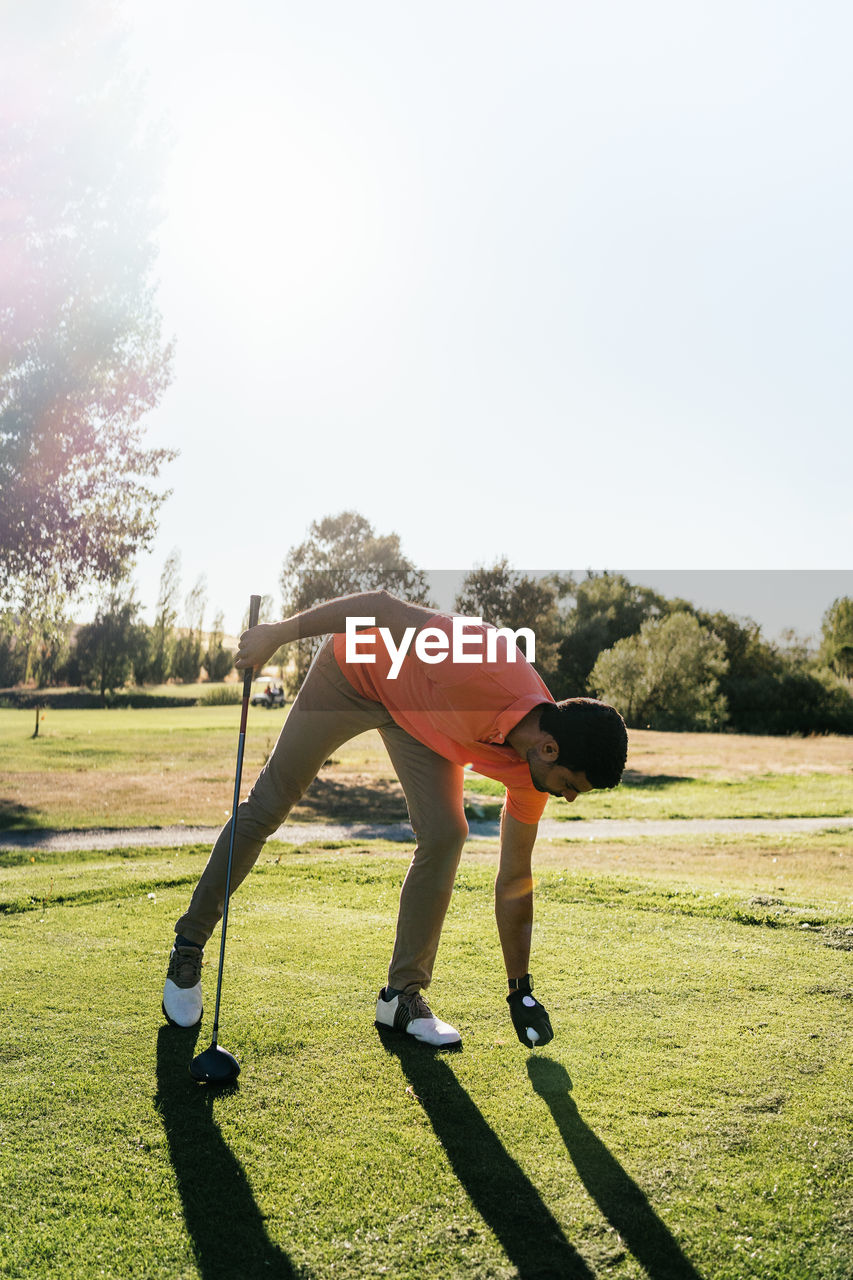 Side view of bearded male golfer leaning forward while putting ball on golf course and looking down in sunlight