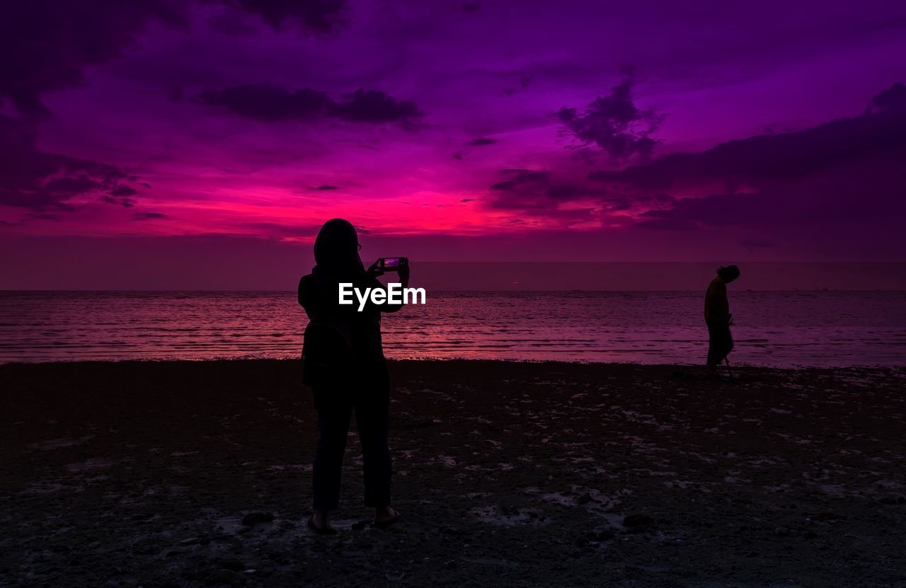 Silhouette woman photographing with mobile phone at beach during sunset