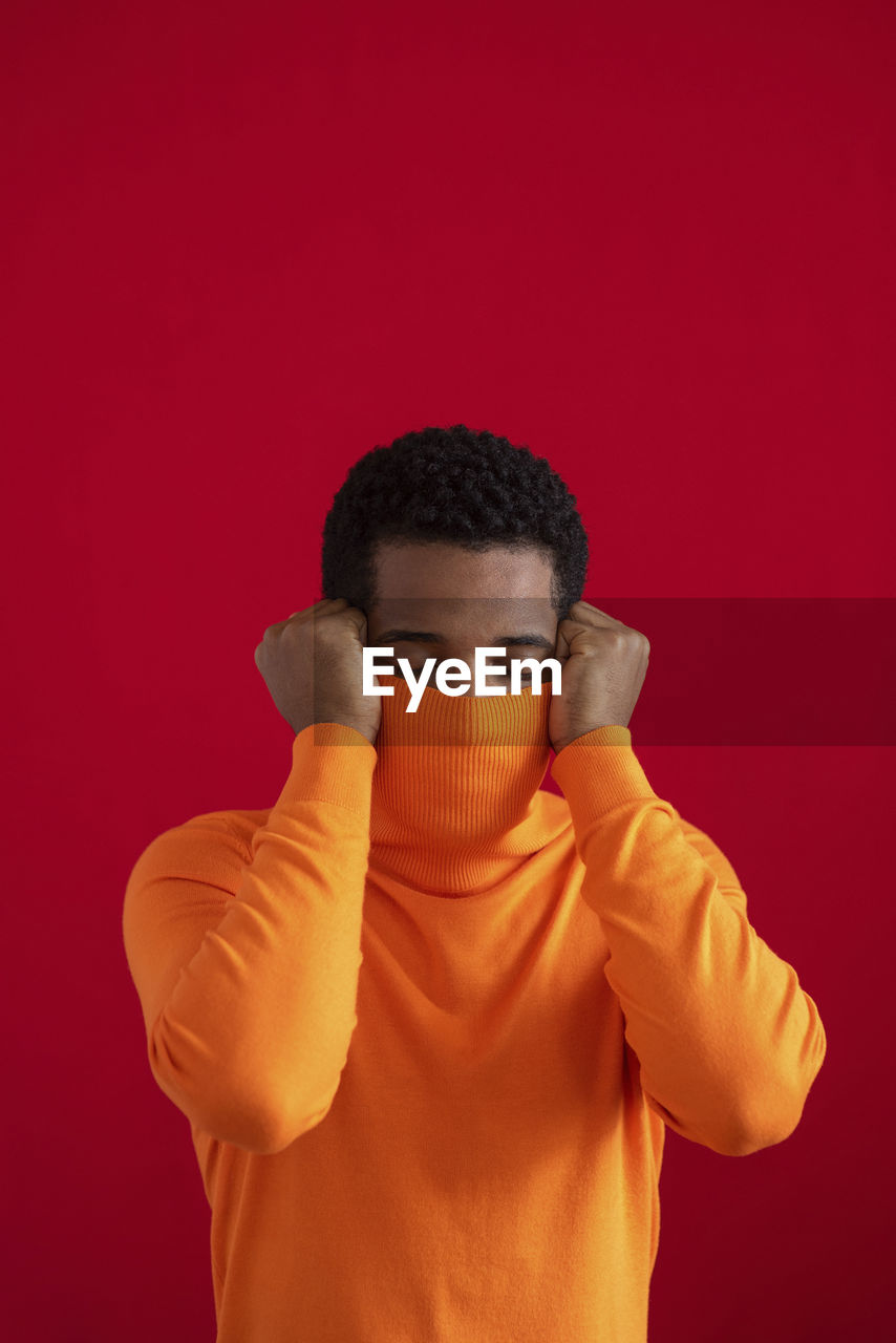 Man covering face with turtleneck sweater against red background