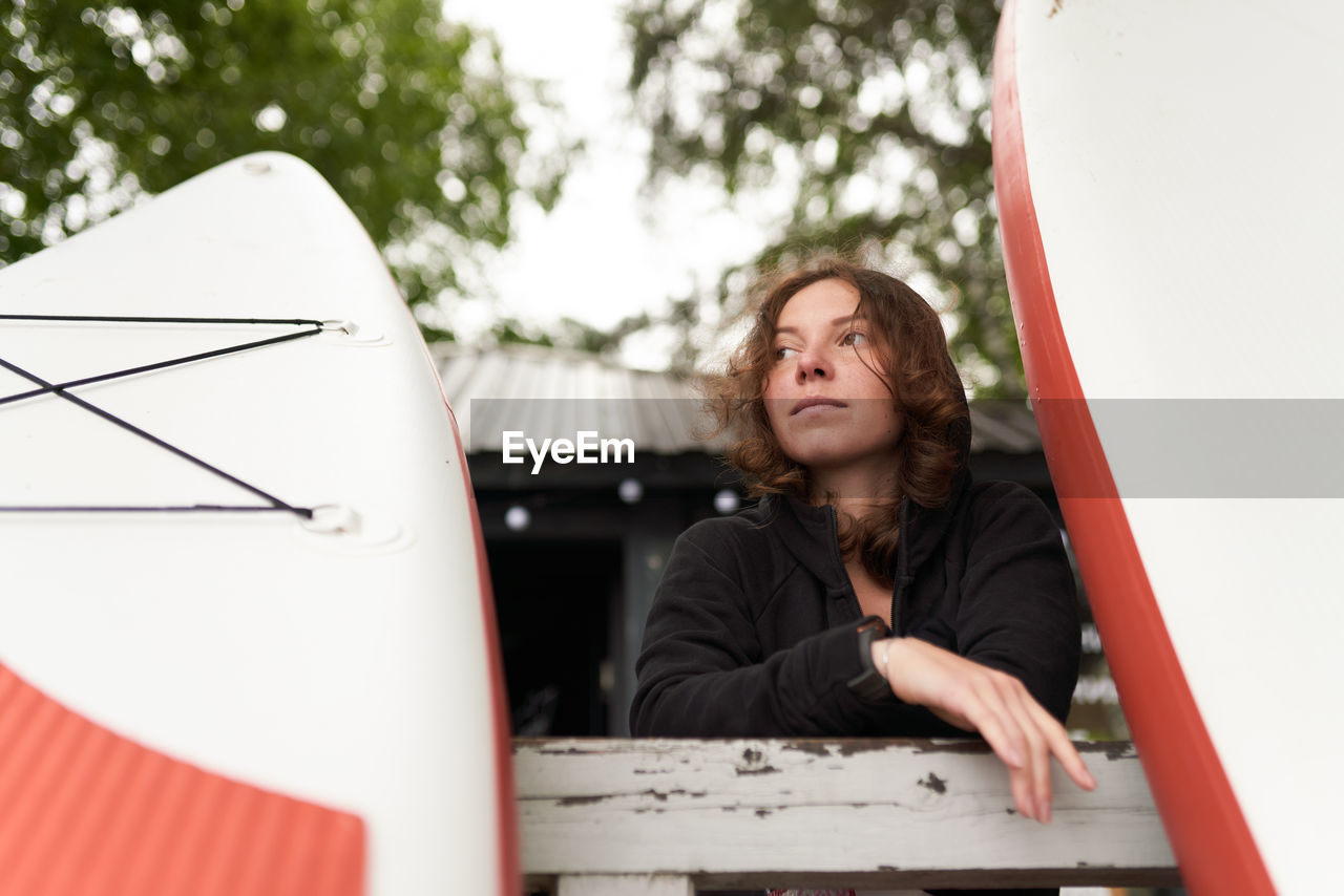 Low angle of calm female surfer stranding on wooden embankment with paddle board while relaxing after training and looking away