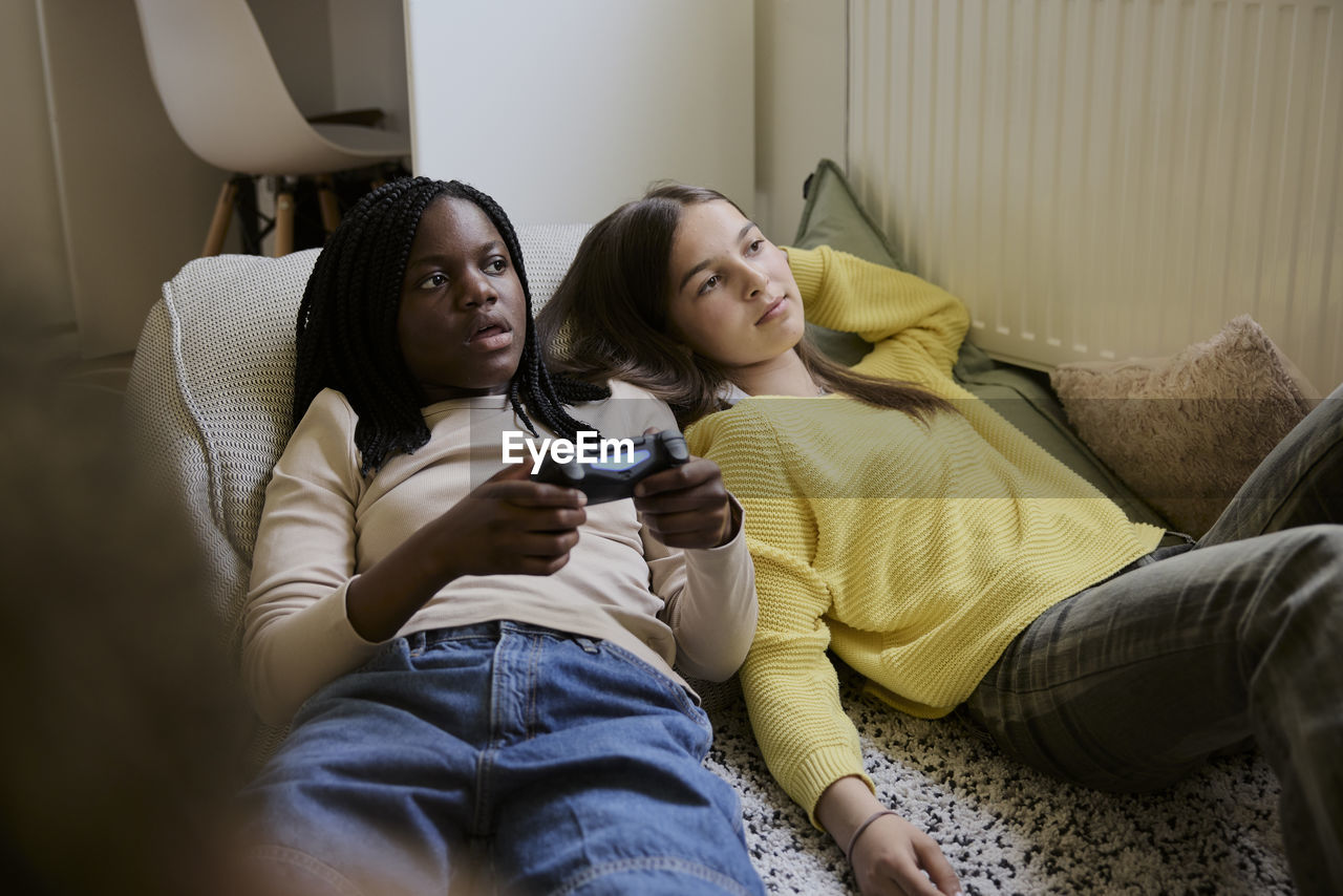 Teenage girl playing video game while lying down with female friend in bed at home