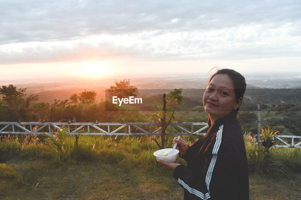 one person, sky, nature, smiling, morning, happiness, women, lifestyles, food and drink, adult, landscape, leisure activity, portrait, young adult, drink, sunlight, environment, plant, beauty in nature, standing, land, cloud, cup, emotion, tranquility, female, relaxation, refreshment, outdoors, sun, child, enjoyment, waist up, rural scene, cheerful, scenics - nature, food, looking at camera, person, field, holding, copy space, tree, drinking, childhood, hairstyle, mug, tranquil scene, side view, day, looking, vacation, lens flare, casual clothing, clothing