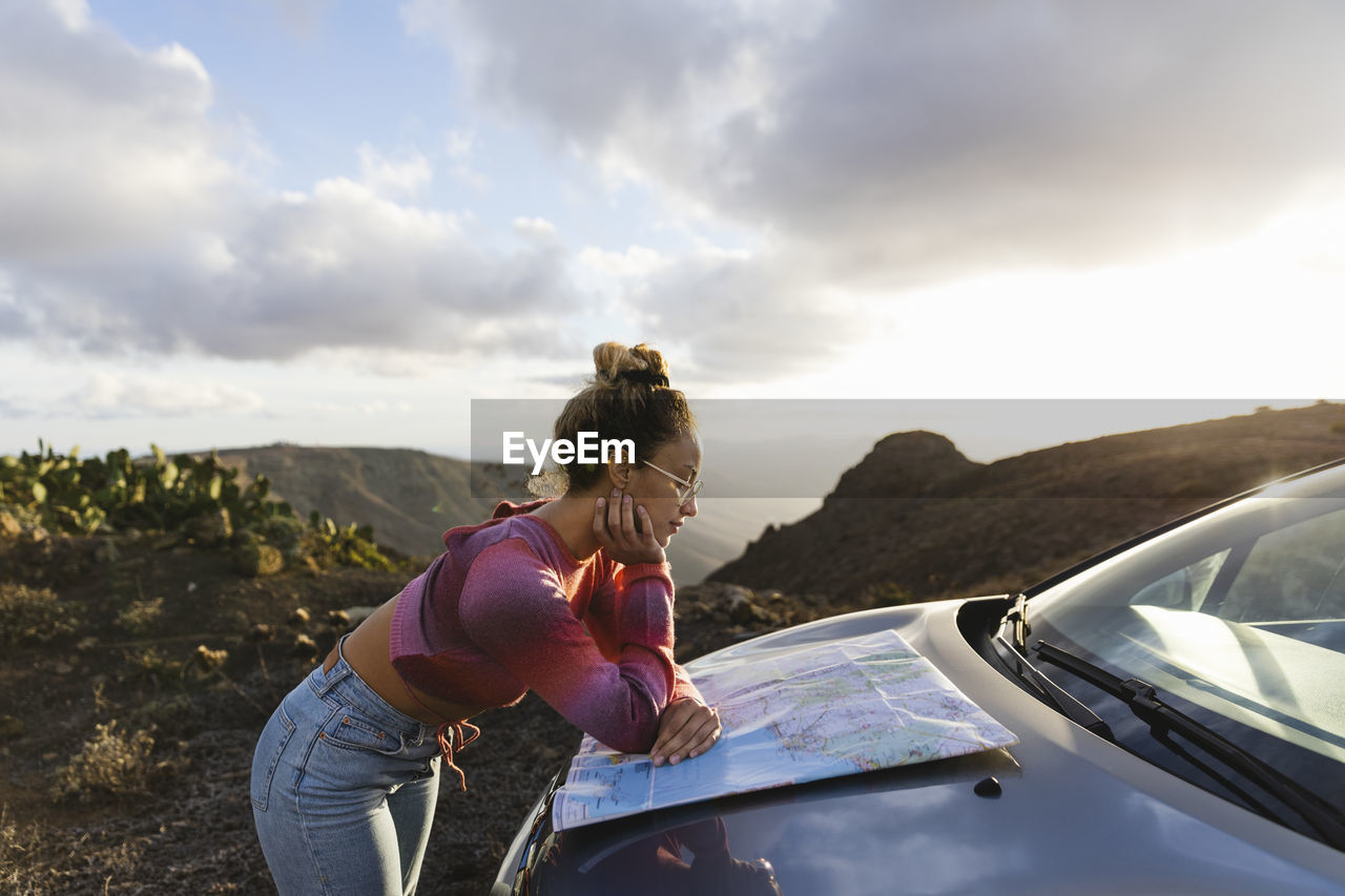 Young woman leaning on car hood checking map at sunset