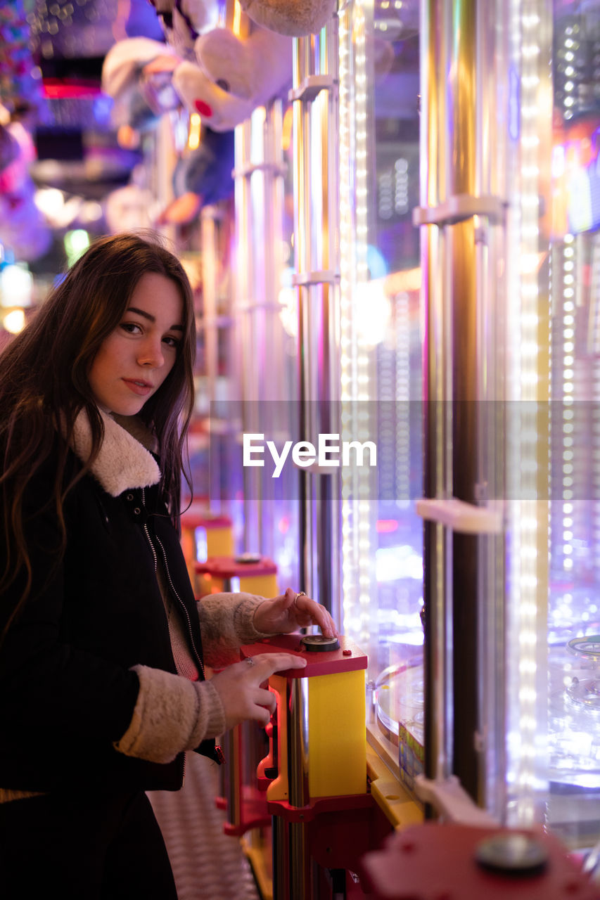 YOUNG WOMAN HOLDING CAMERA WHILE STANDING AT ILLUMINATED ROOM