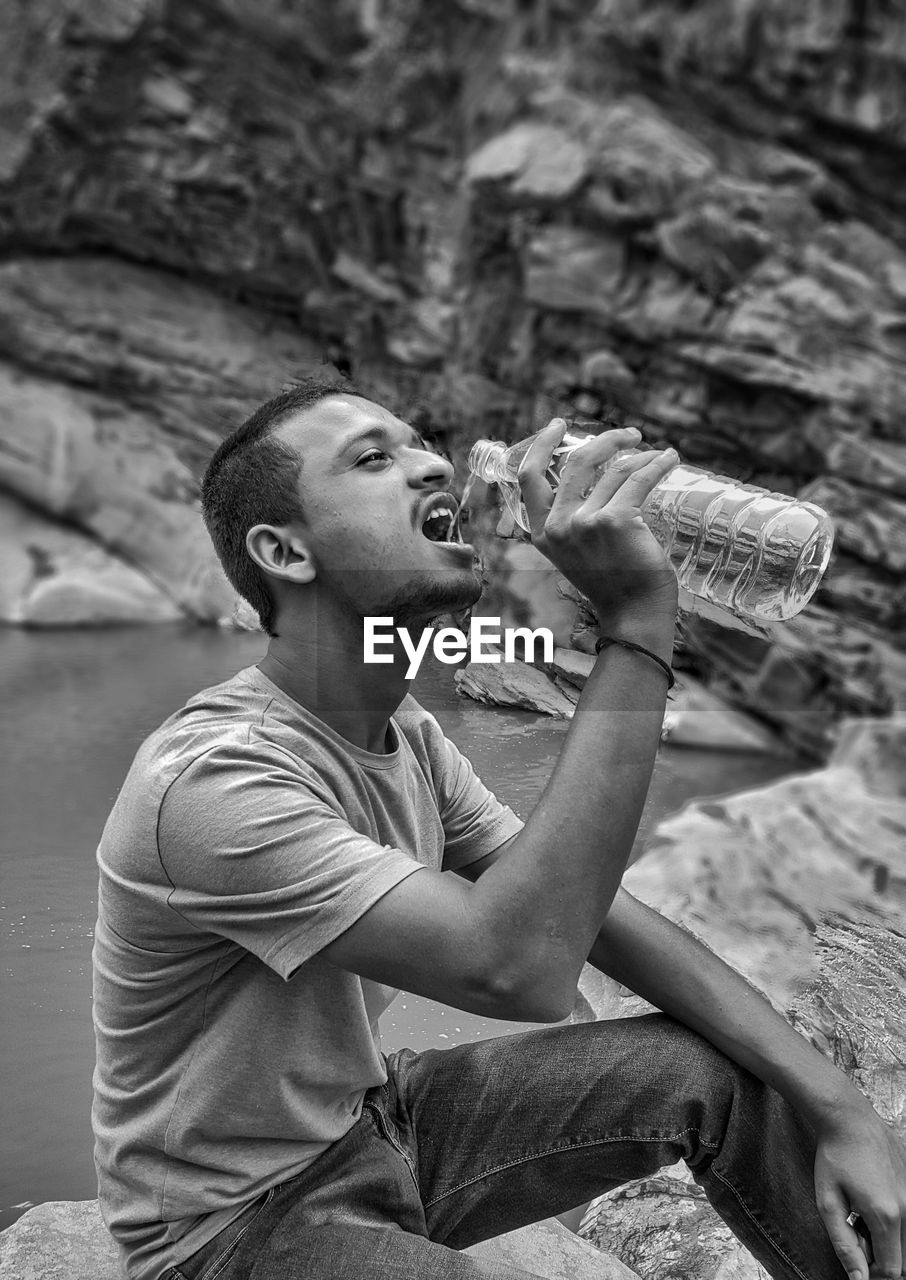 Man drinking water while sitting on rock at riverbank