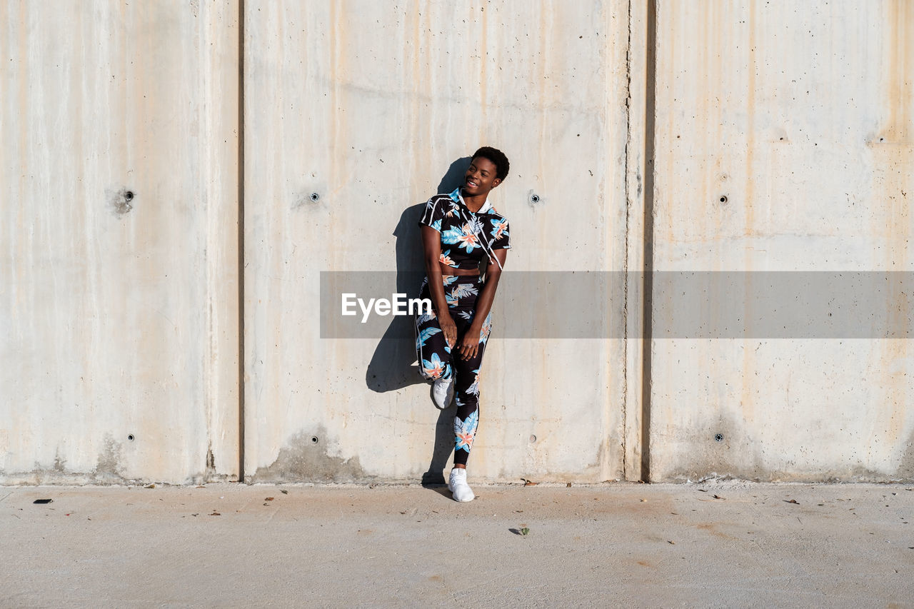 Cheerful confident african american female athlete in flowered sport clothes and white sneakers looking away with interest and laughing while standing alone leaning on concrete wall in sunbeams and resting after training in city