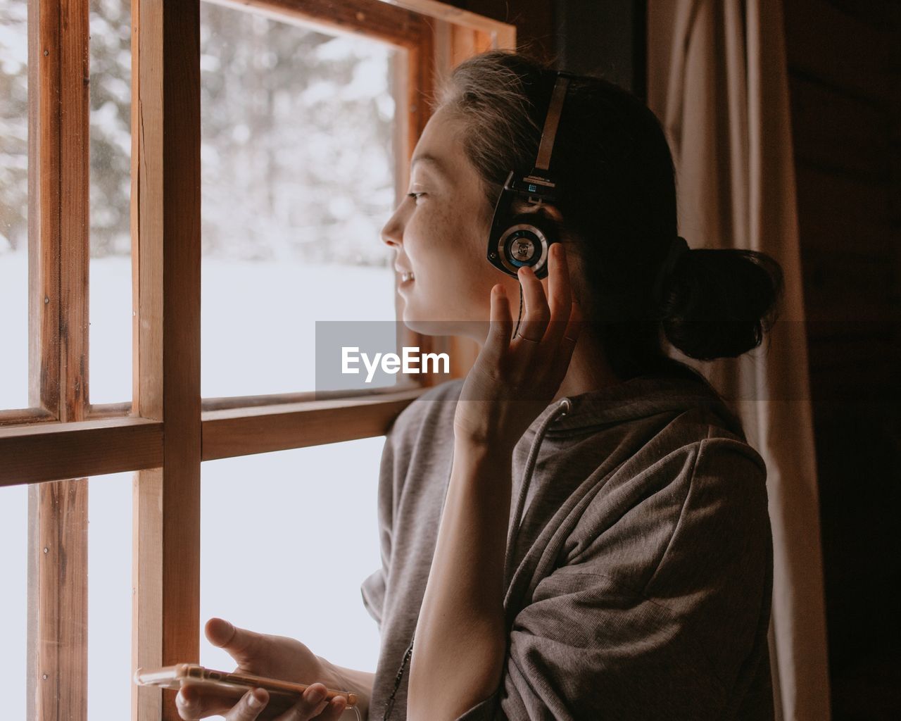 Woman in country house at the window with headphones and mobile phone