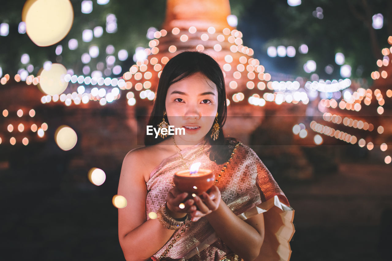 Portrait of young woman holding diya while standing against illuminated temple at night