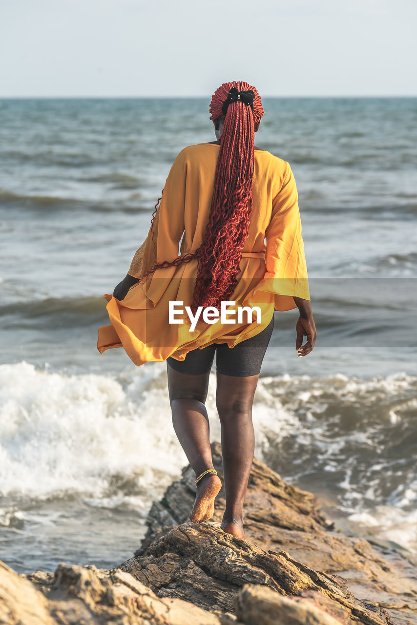 African woman walking on rocks by the beach in accra. has braided red rasta hair, ghana west africa.