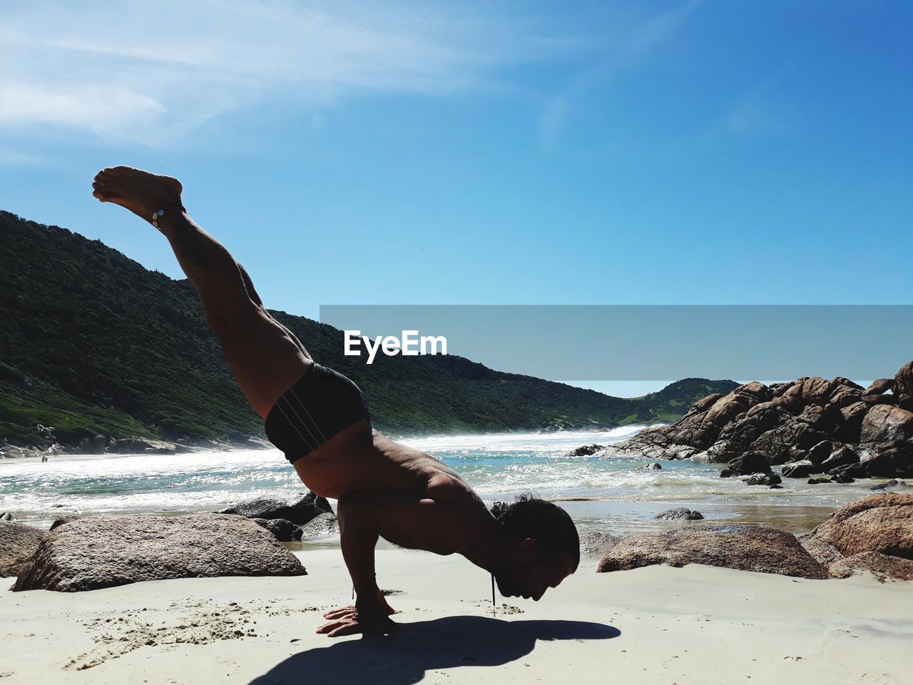 Full length of shirtless man practicing handstand at beach against blue sky