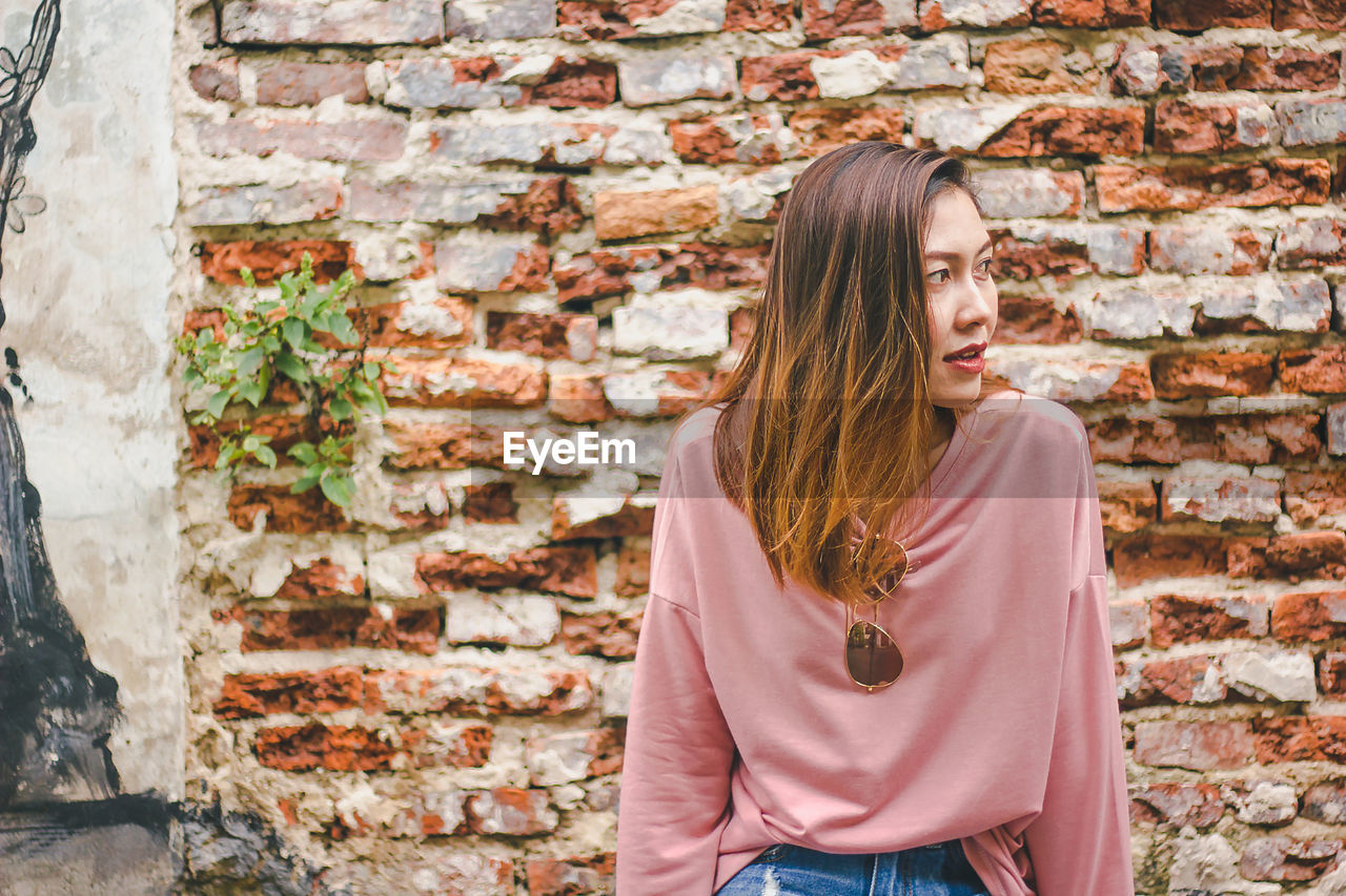 Mid adult woman looking away while sitting against brick wall