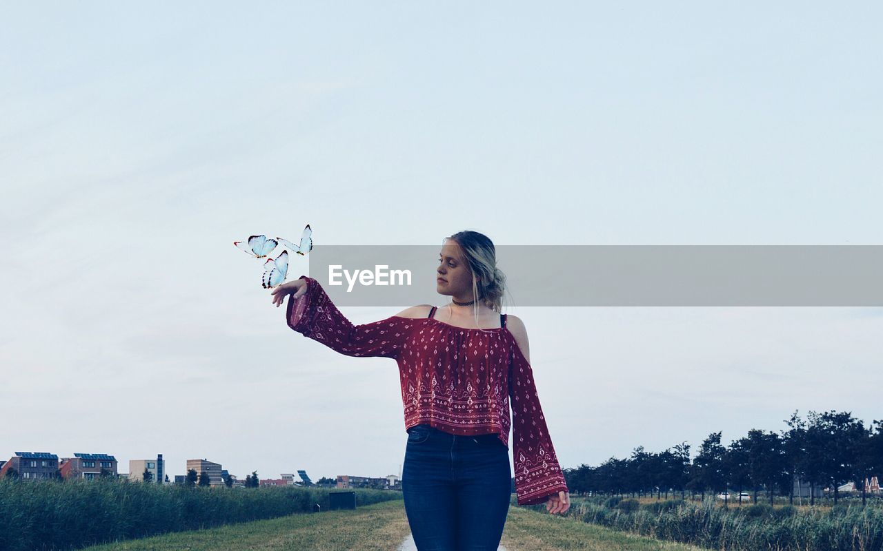 Digital composite image of butterflies flying by woman on field against sky