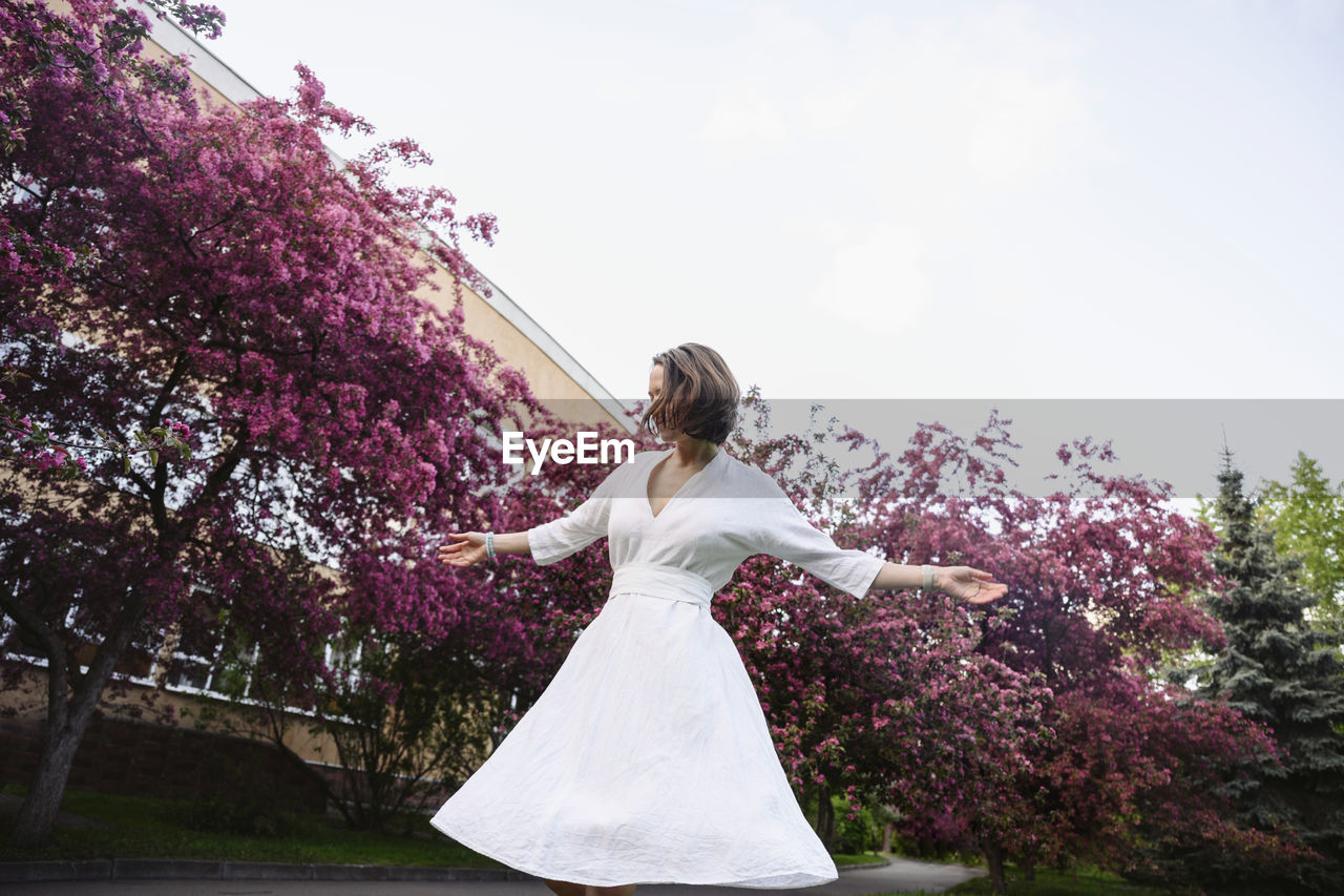 Woman wearing white dress with arms outstretched in apple blossom garden