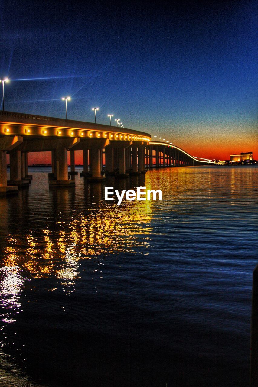 ILLUMINATED BRIDGE OVER RIVER AGAINST SKY AT DUSK