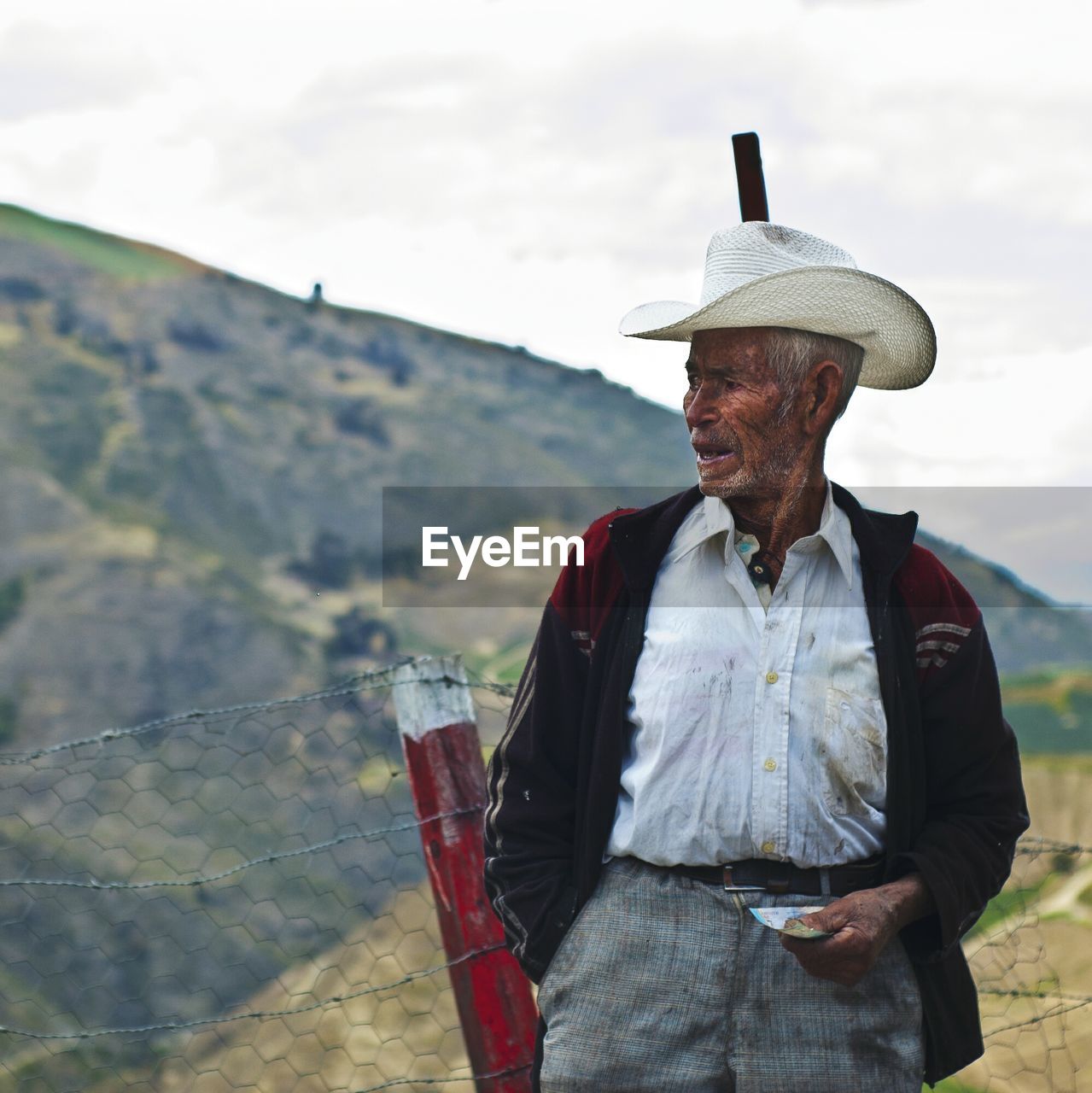 Senior man wearing cowboy hat standing by fence against sky