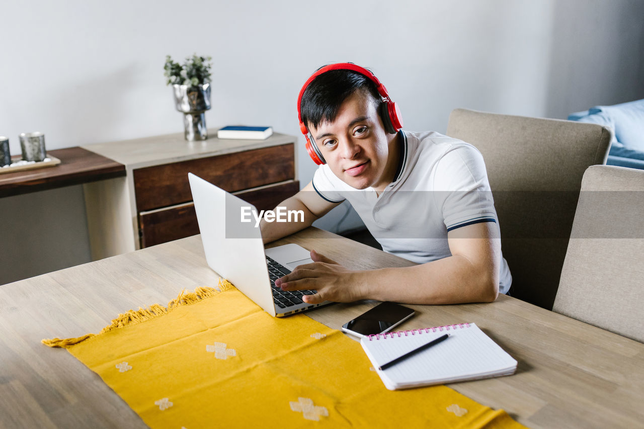 High angle of content latin teen boy with down syndrome browsing netbook while sitting at table and studying online from home