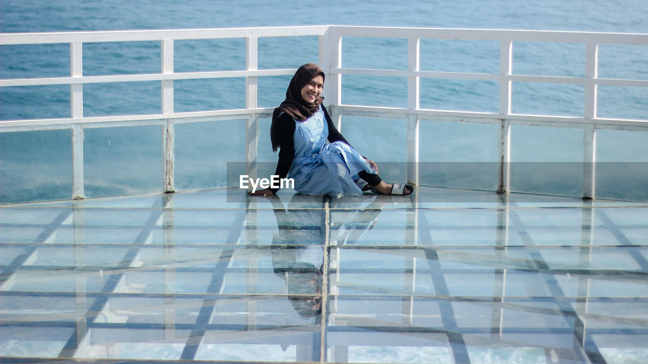 Portrait of smiling woman sitting by railing against sea