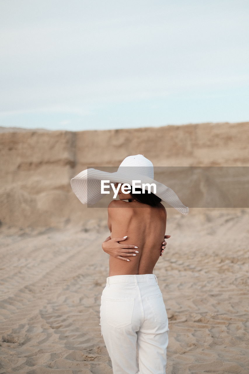 Rear view of shirtless woman wearing hat standing against sand dune