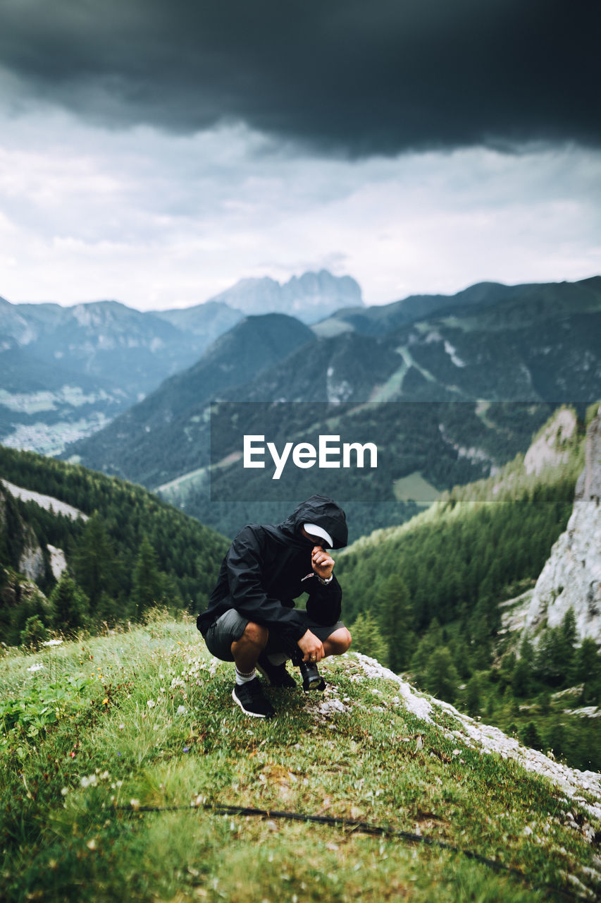 Man wearing hoodie crouching against mountain range