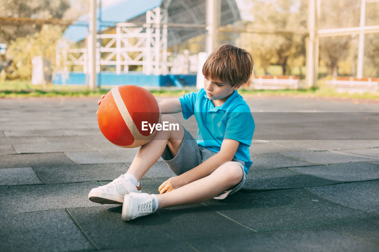 A boy basketball player in a blue t-shirt is sitting on the basketball court with a ball in his hand