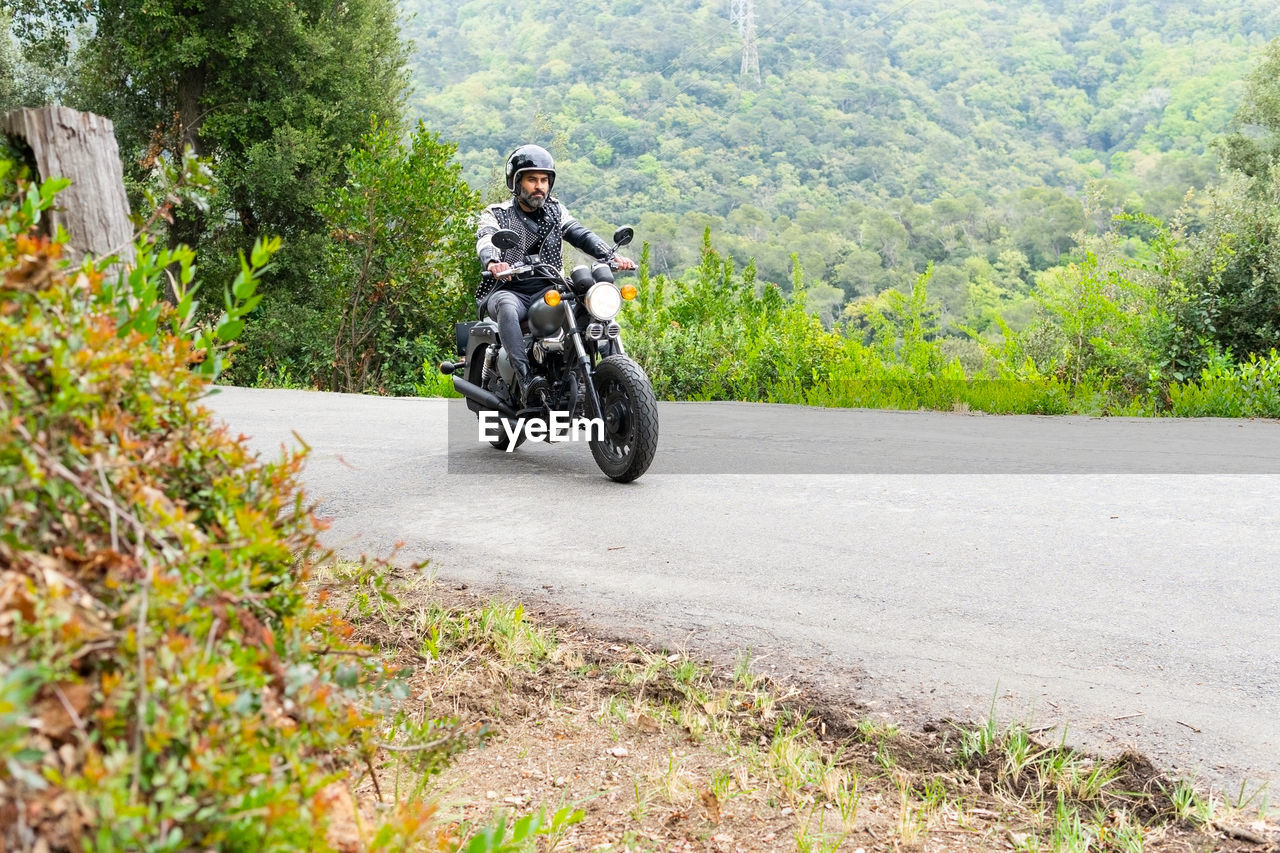 Full body of focused bearded ethnic male biker in black leather jacket and helmet riding modern motorbike on asphalt road amidst lush green trees growing in mountainous valley