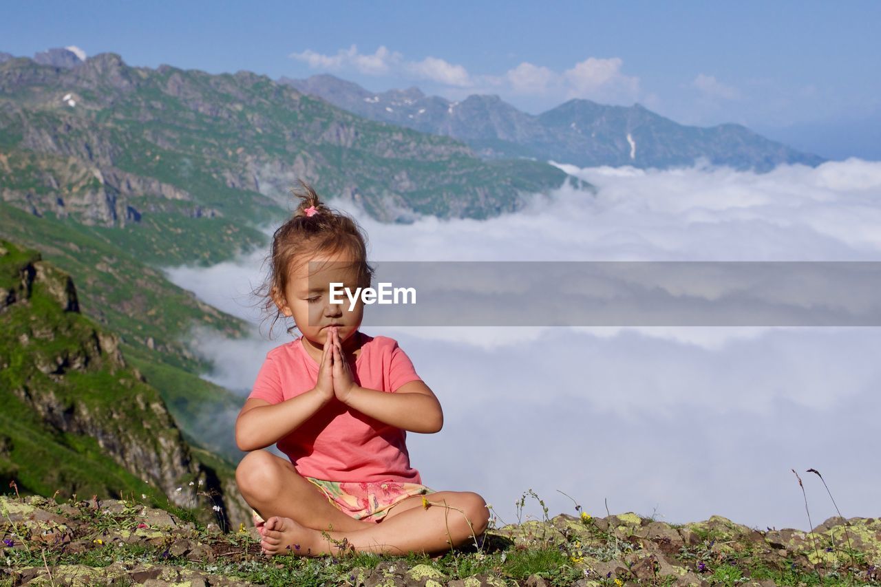 Girl meditating while sitting on mountain against sky