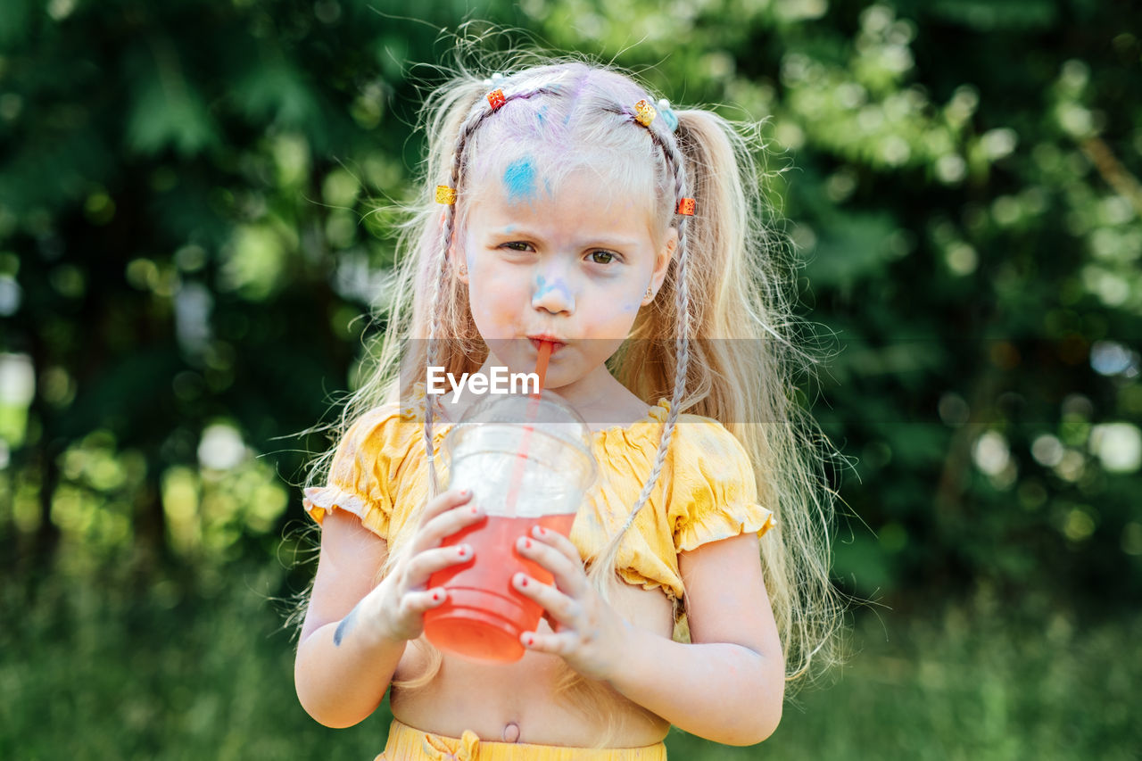 Outdoor portrait of funny grimy little girl with two ponytails drinks lemonade sweet sweetened