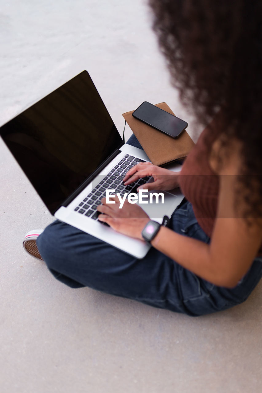 African american self employed woman sitting on street with laptop and browsing smartphone