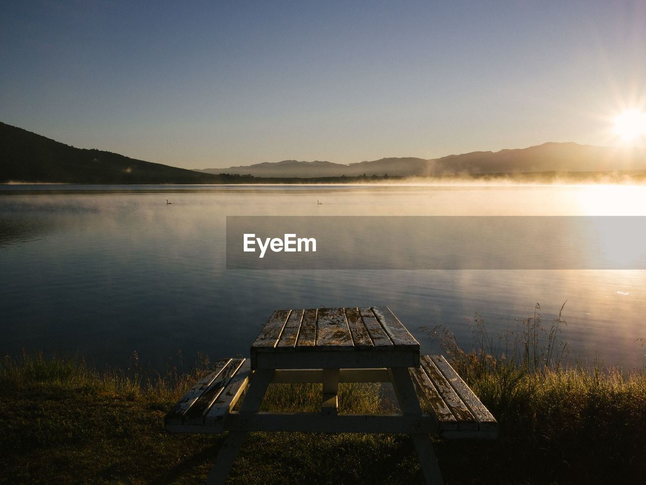 Picnic table at lakeshore against sky during sunrise