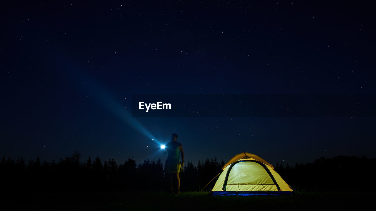 Full length of man flashing light in sky while standing by tent at night