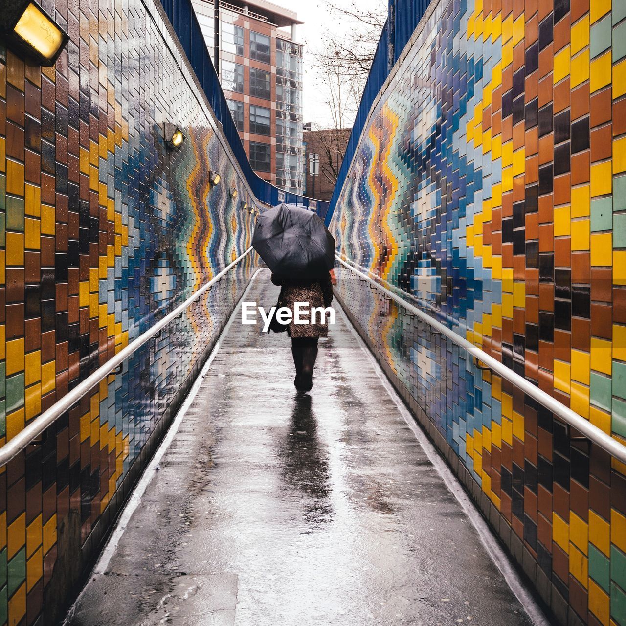 Rear view of woman under umbrella walking on footpath amidst colorful wall