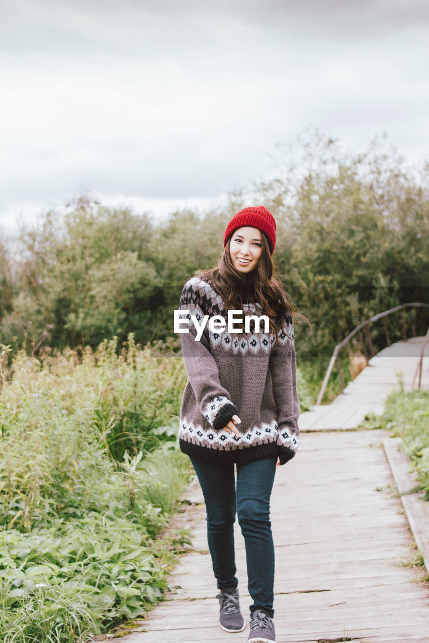 Portrait of smiling young woman walking on footpath against trees and plants