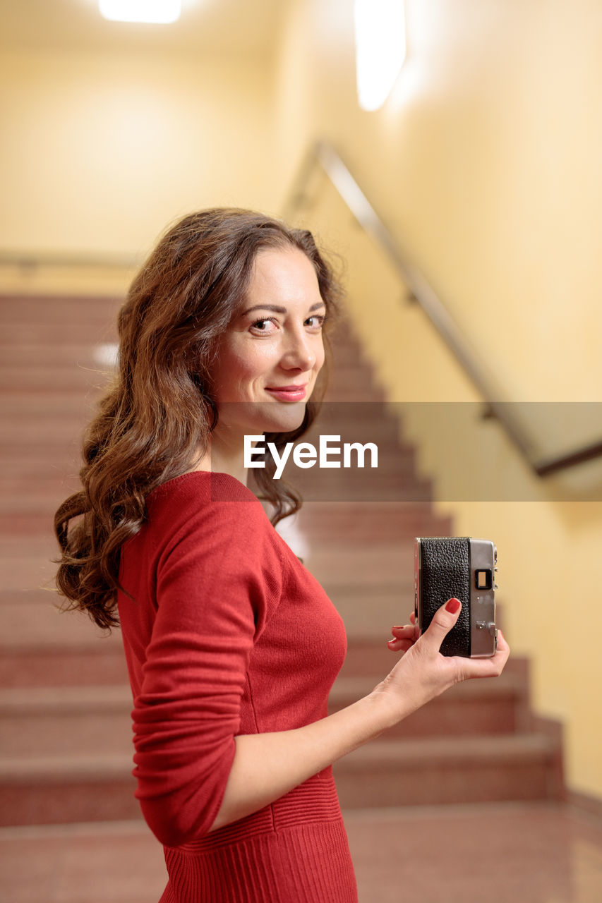 Portrait of young woman with camera against staircase