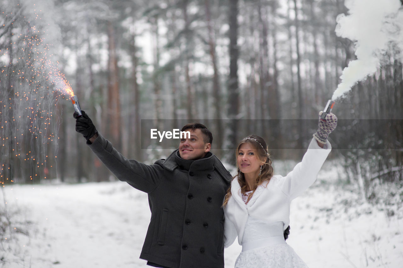 Smiling wedding couple holding firework and distress flare while standing at forest during winter